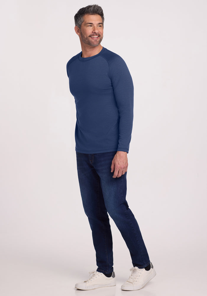 A man with short dark hair and a beard is wearing the Woolx Essential Tee - Starry Night, dark jeans, and white sneakers. He stands smiling against a plain light background. The temperature-regulating tee keeps him comfortable.