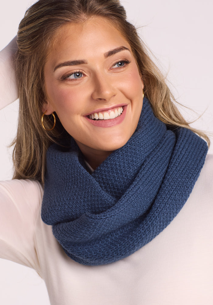 A smiling woman wears the Woolx Noelle Infinity Scarf in Starry Night, paired with a white long-sleeve top and gold hoop earrings, her hand resting on her head against a light background—showcasing this chic winter accessory.