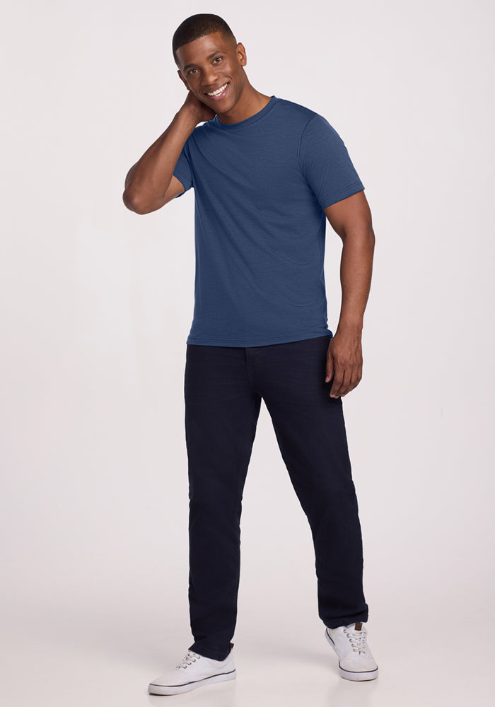 A smiling man stands against a plain light background, wearing the Woolx Endurance Tee in Starry Night, paired with dark pants and white sneakers. One hand touches the back of his neck while the other rests by his side.