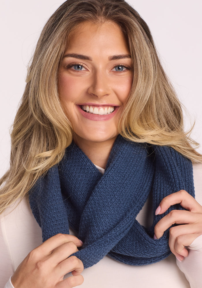 A smiling woman with long, wavy blonde hair models the Woolx Noelle Infinity Scarf in Starry Night, pairing the textured, dark blue merino wool accessory with a light top and holding it against a plain background.