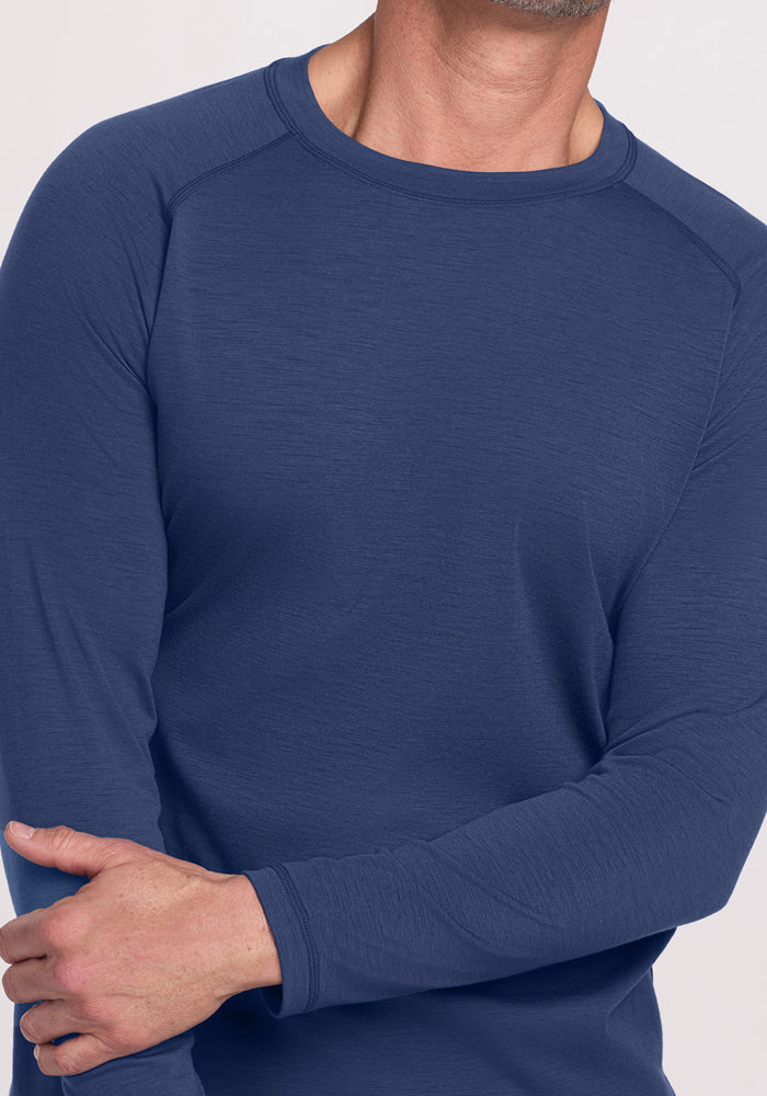 A man models the Woolx Essential Tee - Starry Night, a lightweight blue athletic shirt, standing with his right arm crossed over to hold his left elbow. His face is mostly out of frame.