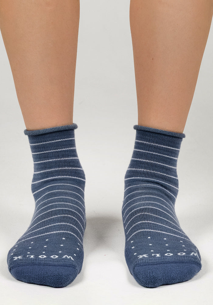 A pair of feet wearing Woolx Womens Derby Roll Top Socks in Vintage Indigo with thin white stripes stand on a light gray surface. These comfy socks have non-slip dots and text on the soles.