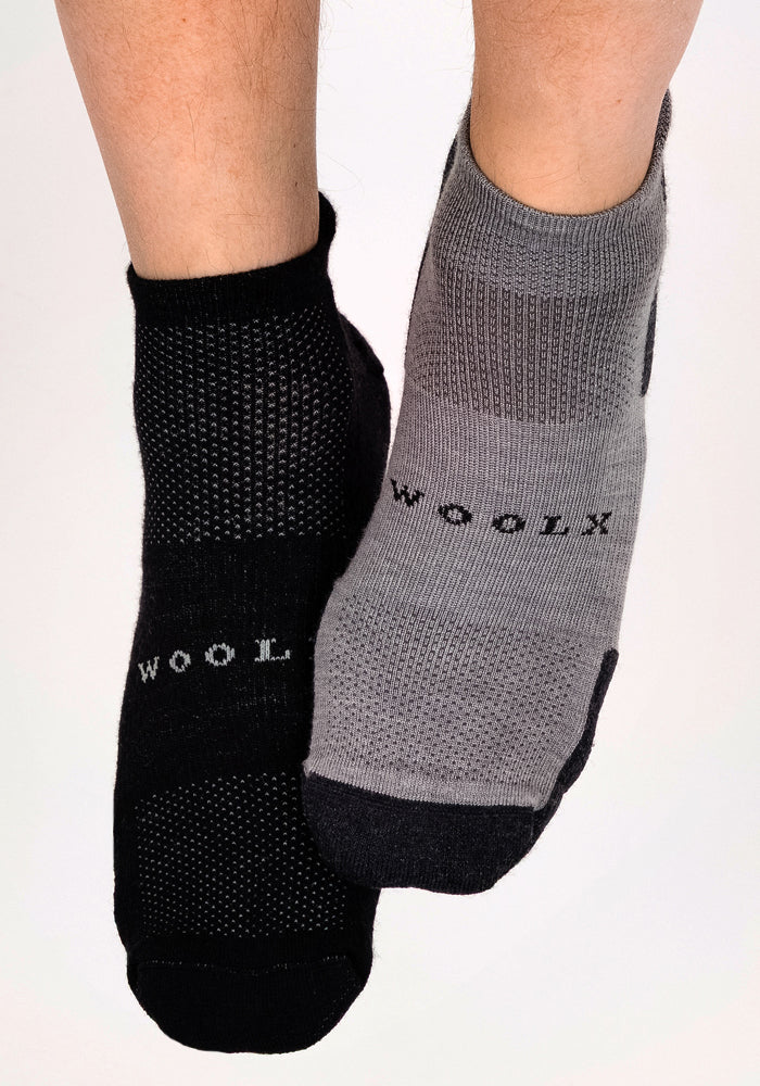 Two feet wear mismatched Woolx Mens Rowan No Show Socks in Pebble; the left foot has a black sock, and the right a light gray sock with black accents. Both textured, moisture-wicking socks feature "Woolx" on top.