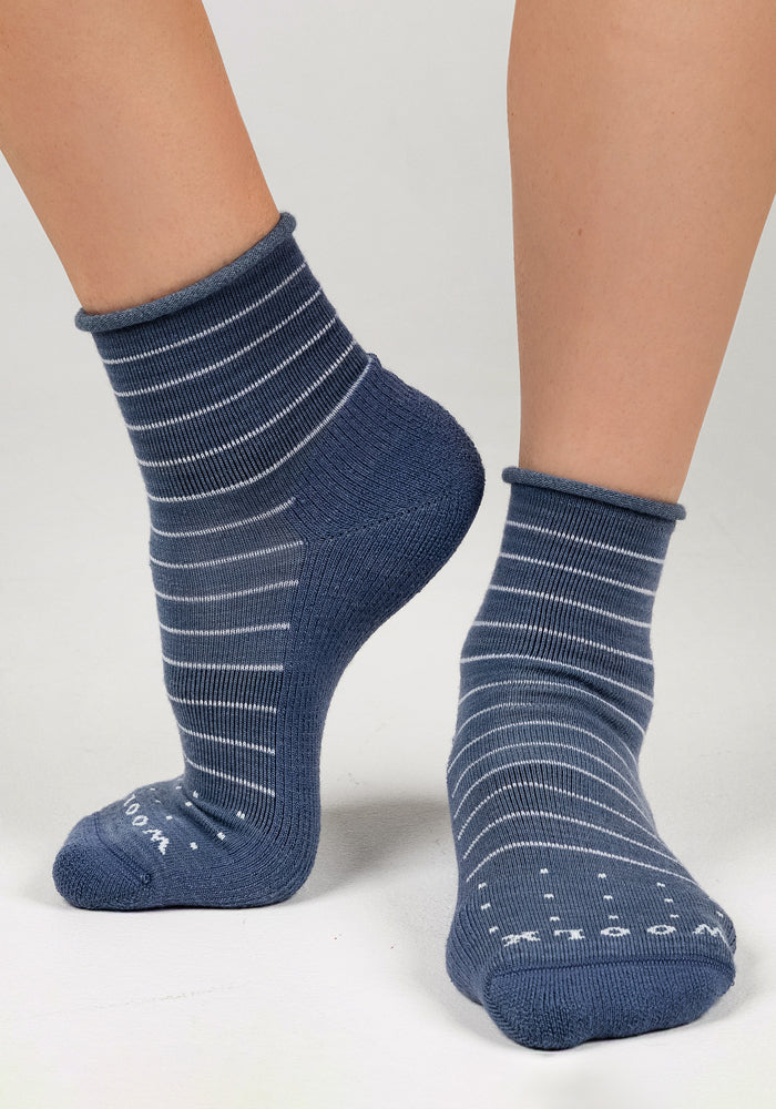A close-up of two feet in Woolx Womens Derby Roll Top Sock - Vintage Indigo, featuring blue with white stripes, grip dots on the soles for stability and comfort, standing on a light gray surface.