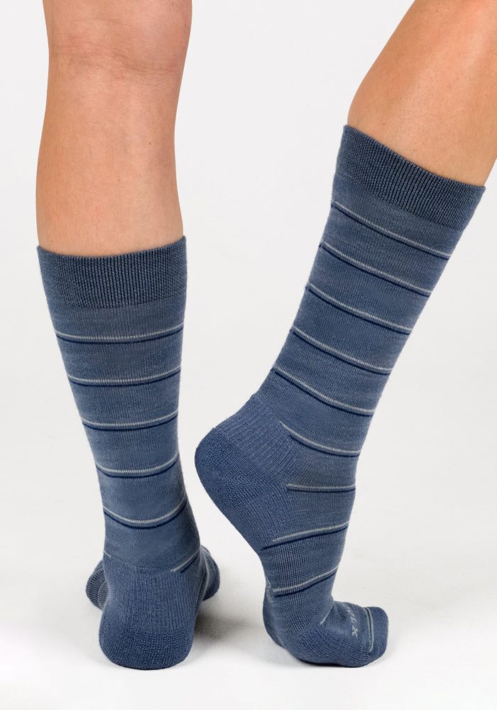 A person stands on a white background wearing Woolx Womens Chandler Mid Calf Socks in Vintage Indigo, which feature thin light-colored horizontal stripes. The socks cover the calves; one foot is flat while the other heel is slightly raised.