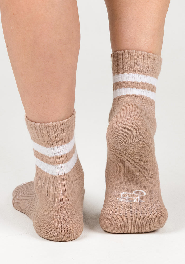 Close-up of two feet wearing Woolx Womens Varsity Mini Crew Socks in Morning Latte, featuring two white stripes near the top and a small white logo on the sole. Background is plain and light-colored.