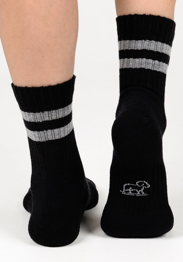 A close-up of two feet wearing Woolx Womens Varsity Mini Crew Socks - Black Volt, featuring two gray stripes near the top and a small white embroidered animal on the sole of the right sock.