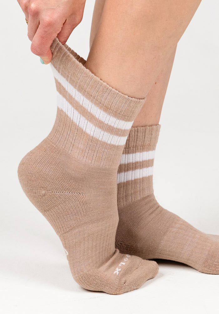 A close-up of a person pulling up the Woolx Womens Varsity Mini Crew Socks in Morning Latte, featuring two white stripes and a matching sock on the other foot against a plain white background.