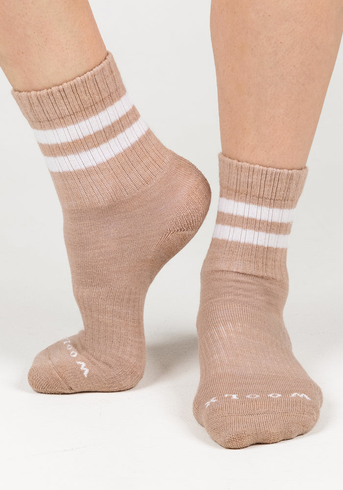A pair of feet in Woolx Womens Varsity Mini Crew Socks - Morning Latte stand on a white surface. The light brown socks have two white stripes near the ribbed cuffs, moisture-wicking fabric, and white text on the toes.