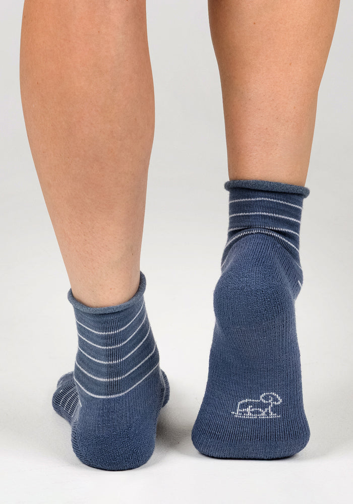 A close-up of a person's lower legs and feet wearing mismatched Woolx Womens Derby Roll Top Socks in Vintage Indigo; the left sock features white stripes, while the right is solid blue with a white logo on the heel.