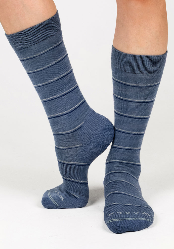 A person wearing Woolx Womens Chandler Mid Calf Socks in Vintage Indigo with thin light gray stripes stands on tiptoe on one foot with the other flat against a plain white background.