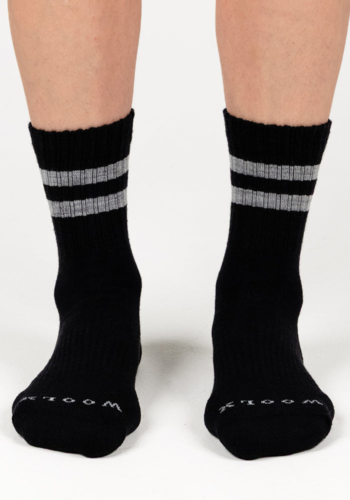 A pair of feet wear Woolx Womens Varsity Mini Crew Socks in Black Volt, featuring two gray stripes near the top and "WOODS" in white on the toes, standing on a white surface.
