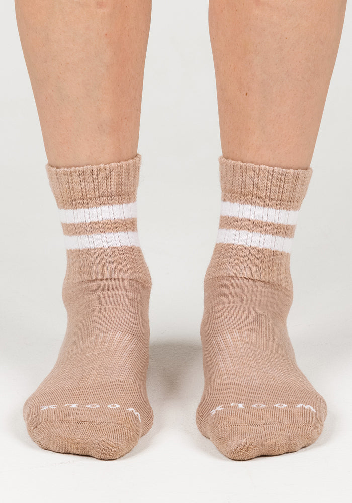 A close-up of two feet wearing Woolx Womens Varsity Mini Crew Socks in Morning Latte, light brown with two white stripes near the top, standing on a white surface.