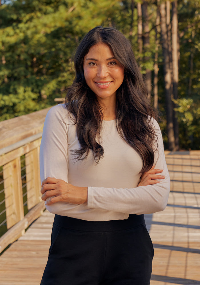 A woman with long dark hair stands on a wooden bridge outdoors, smiling with arms crossed while wearing the Woolx Sophia Bodysuit - Black and black pants. Sunlight and trees highlight the bodysuit's moisture-wicking comfort.