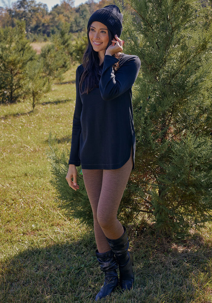 Woman in Woolx Stella Leggings - Sable Heather, black sweater, boots, and beanie stands outside smiling.