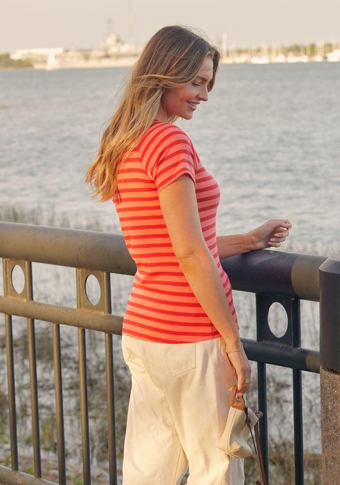 A woman in a Woolx Mia V Neck - Wildberry Stripe tee and white pants stands by a waterfront railing, smiling and looking down, with water and boats in the background.