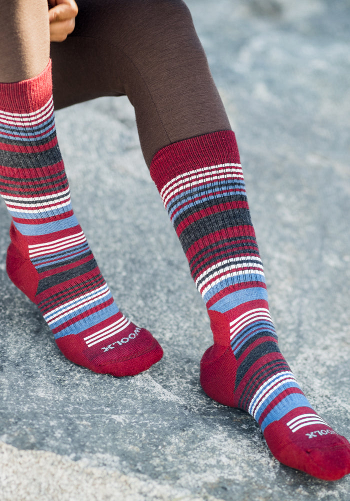 A person in brown leggings wears Woolx Womens Everyday Crew Socks - Rhubarb Stripe, featuring red, blue, white, and black stripes and moisture-wicking cushioning. The Woolx logo appears on the toes as they sit on a gray stone surface.