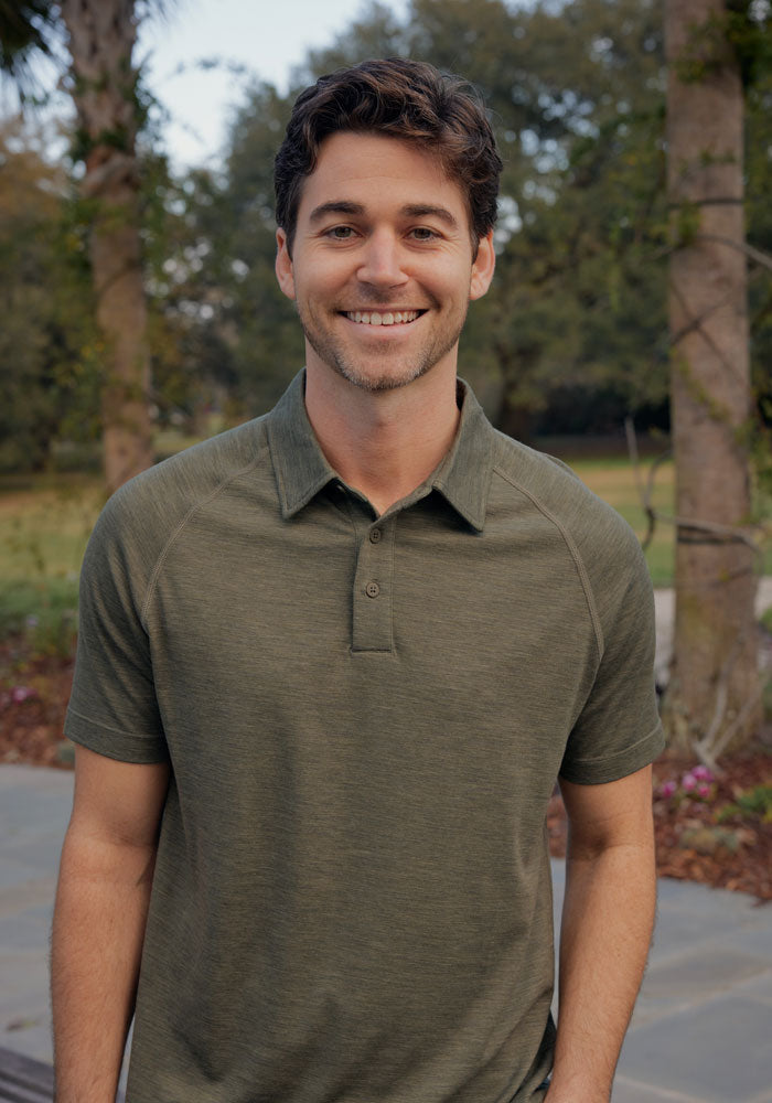 A smiling man with short brown hair wears the Woolx Summit Polo in Sage Olive Melange, standing outdoors on a paved path with trees and greenery, enjoying its moisture management perfect for athletic activities.