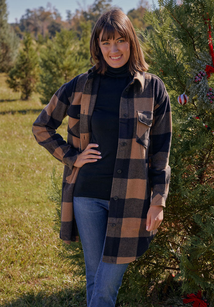 A woman smiles outdoors by a decorated evergreen, wearing the Woolx Sutton Shacket in Camel Plaid over a black turtleneck and blue jeans. The grassy scene has more trees in the background.