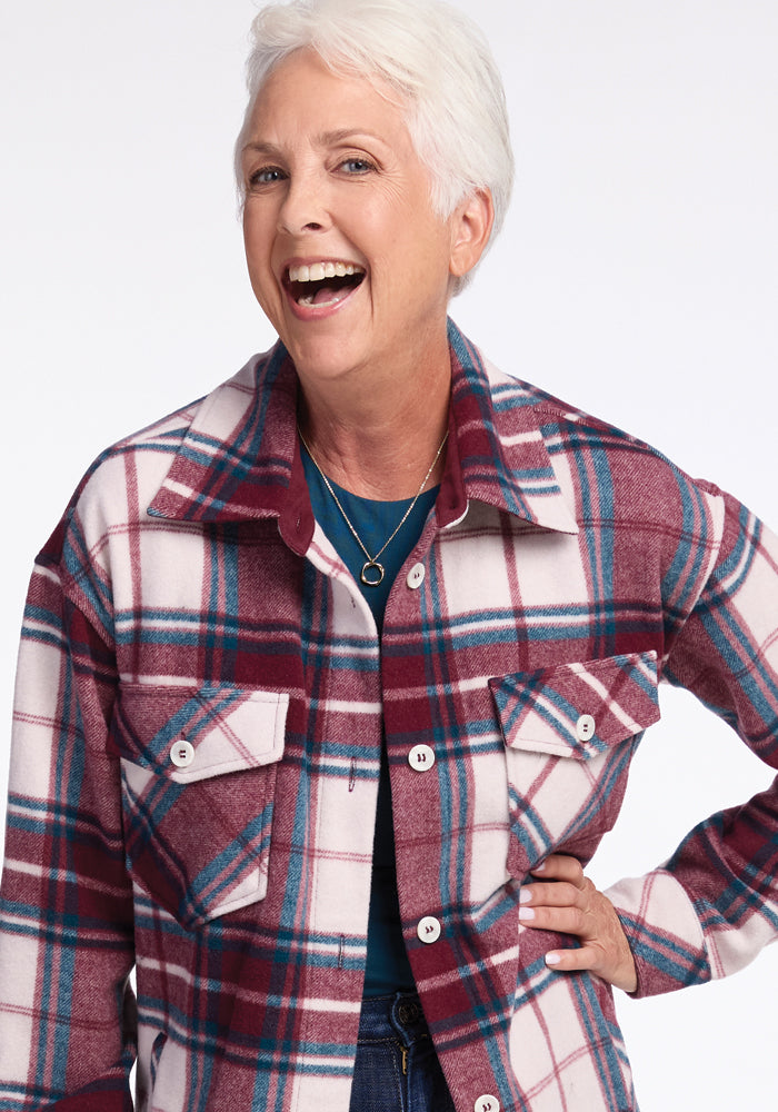 Smiling older woman with short white hair wears the Woolx Sutton Shacket in Frozen Cranberry Plaid over a blue top, standing with one hand on her hip against a plain white background.