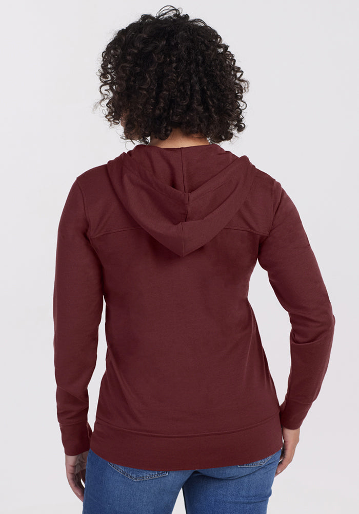 A person with curly hair, seen from the back, wears the Woolx Ryann Hoodie in Toasted Garnet and blue jeans against a plain light background.