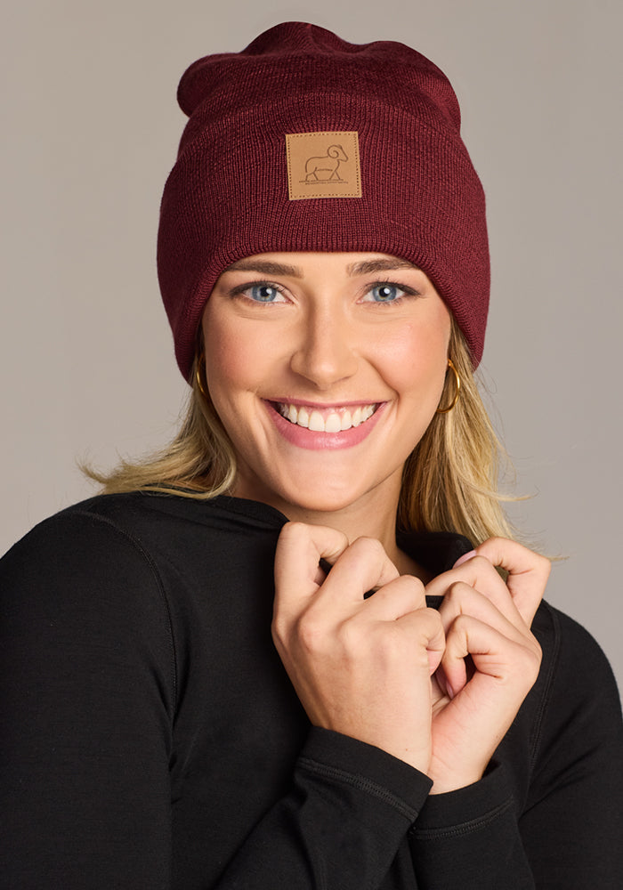 A smiling woman in a black top wears the Woolx Baylor Beanie - Toasted Garnet, featuring a brown patch with a line drawing. Made from soft Australian merino wool, it’s the perfect cozy winter hat.