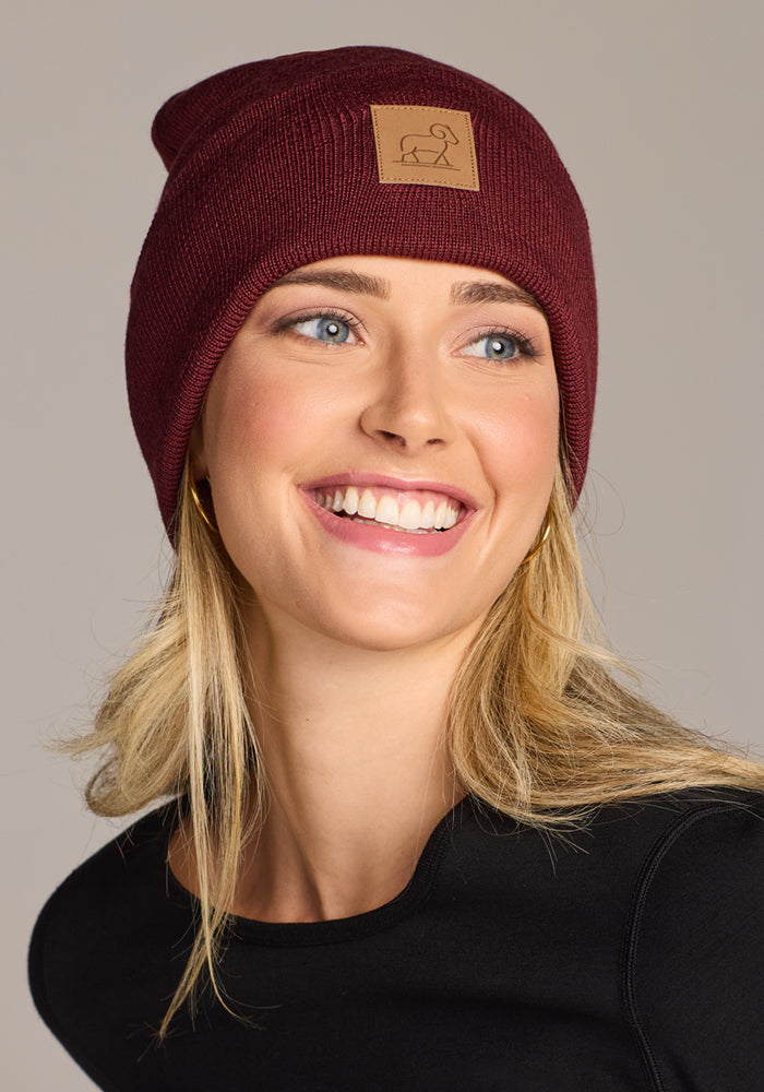 A smiling woman with blonde hair wears the Woolx Baylor Beanie in Toasted Garnet—a maroon merino wool hat with a tan patch and simple line drawing—paired with a black top against a neutral background.