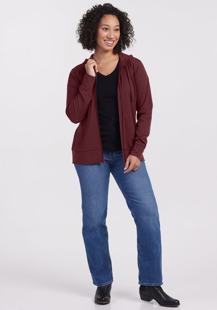 A woman with curly hair smiles in the Woolx Ryann Hoodie in Toasted Garnet over a black shirt, blue jeans, and black ankle boots, standing against a plain white background.