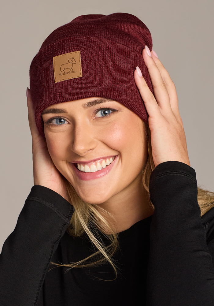 A smiling woman with blonde hair and blue eyes wears the Woolx Baylor Beanie in Toasted Garnet, a warm merino wool hat with a small brown patch featuring a minimalist animal design. She touches the sides of the beanie with both hands.