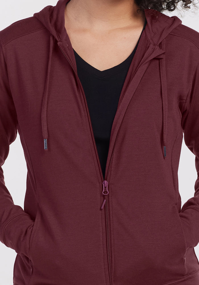 A person wears the Woolx Ryann Hoodie in Toasted Garnet over a black V-neck shirt, hands in pockets. The photo is cropped from neck to mid-hips, highlighting the upper body and hoodie details.