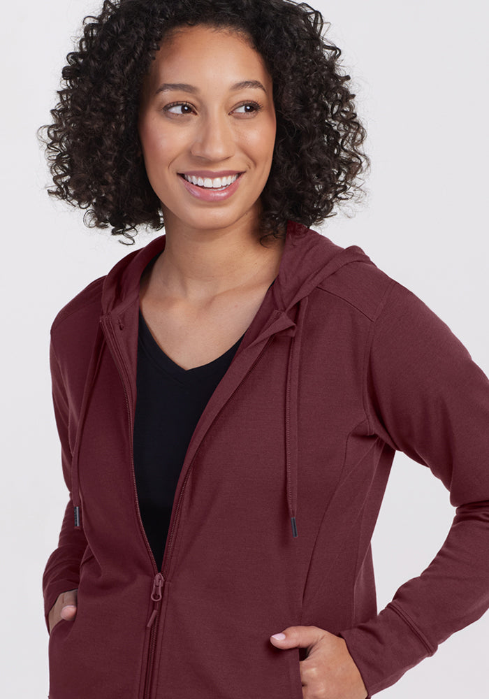 A woman with curly hair smiles while looking to the side, wearing the Woolx Ryann Hoodie in Toasted Garnet over a black top, her hands tucked in the pockets as she stands against a plain light background.