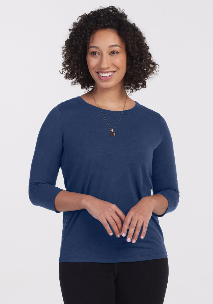 A woman with curly hair smiles while wearing the Woolx Jenny 3/4 Sleeve Crew Neck in Starry Night and black pants, standing against a plain white background. 