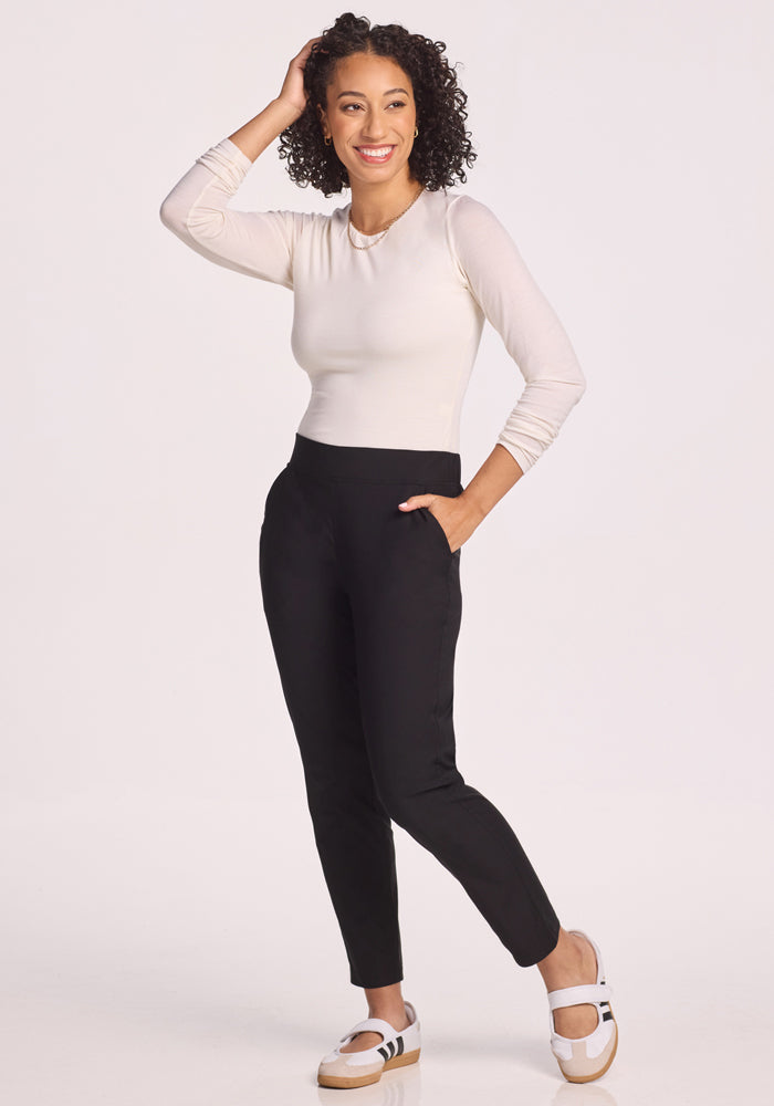 A woman with curly hair smiles, standing with one hand in her pocket and the other touching her head. She wears a long-sleeve white top, Woolx Nicole Pants in black, and white slip-on shoes with black stripes.