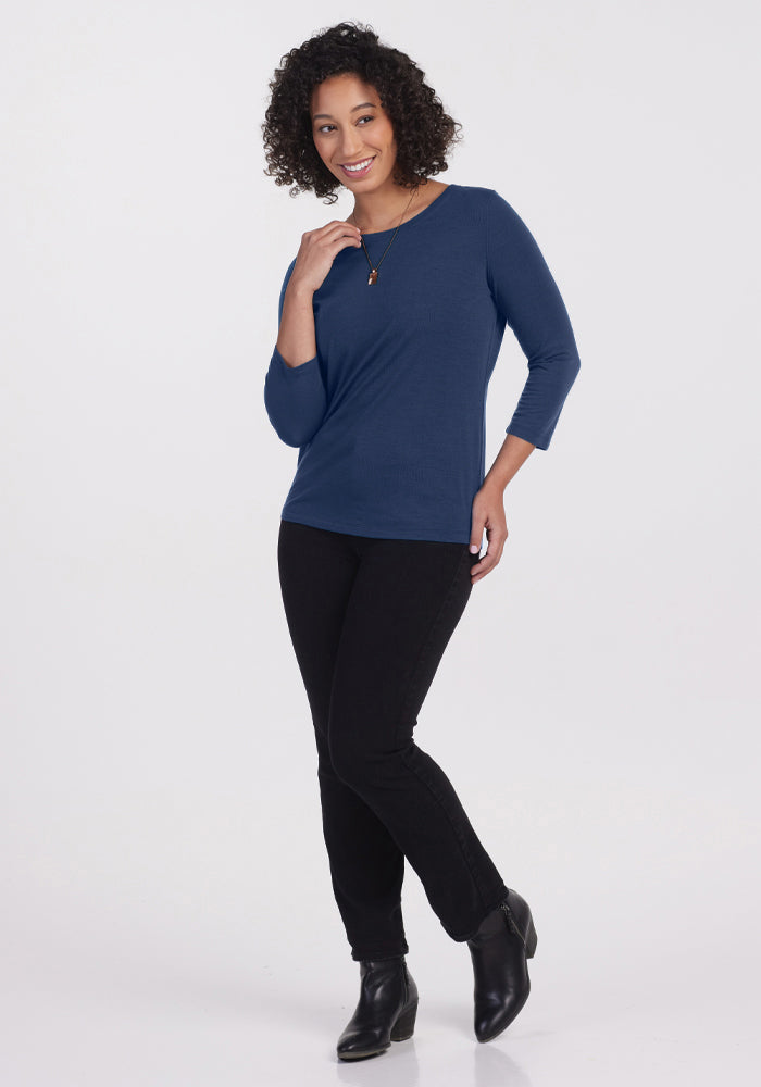 A woman with curly hair smiles while standing, wearing the Woolx Jenny 3/4 Sleeve Crew Neck in Starry Night, paired with black pants, black ankle boots, and a necklace against a plain white background.