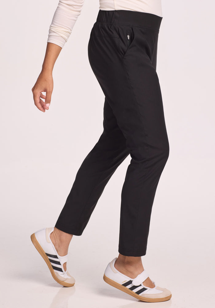 A person in Woolx Nicole Pants - Black walks against a plain light background, wearing breathable black pants, an off-white long-sleeve top, and white slip-on shoes with black stripes; only the lower body is visible.