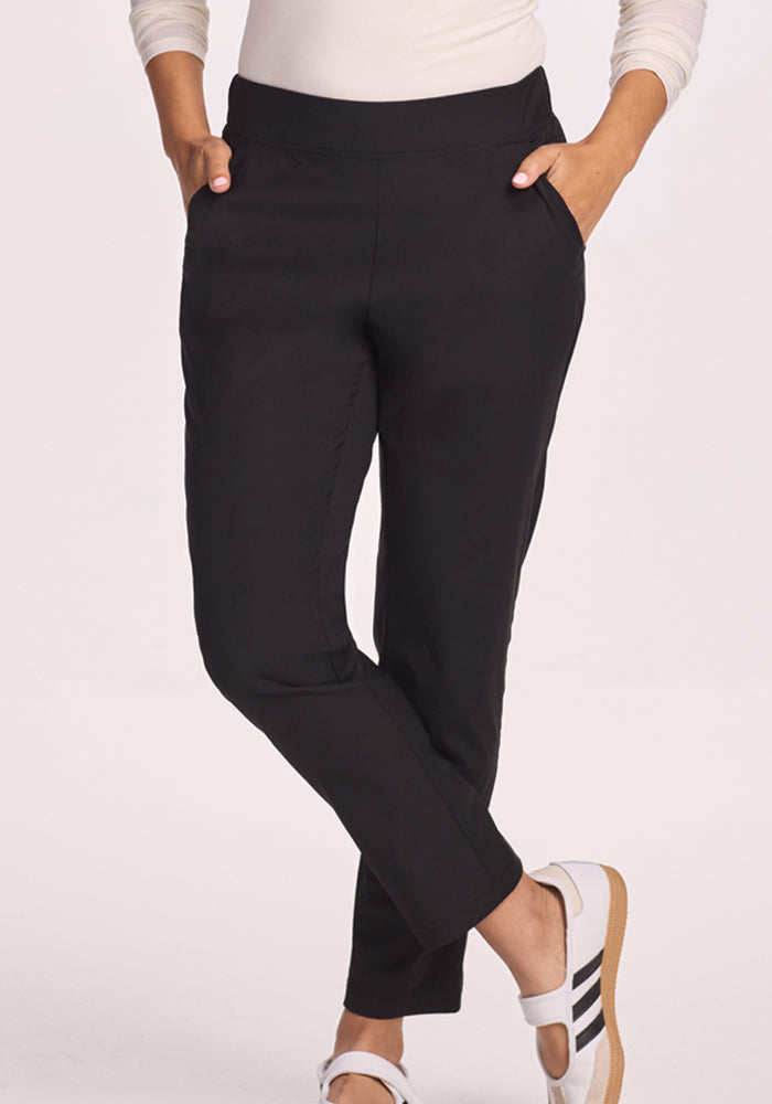 A person wearing Woolx Nicole Pants in black, with hands in pockets, a white long-sleeve top, and white shoes with black stripes, stands cross-legged against a light background.