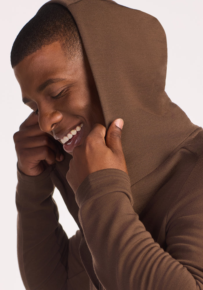 A smiling man with closed eyes pulls up the hood of his Woolx Boulder Hoodie in Bourbon Oak, looking relaxed and happy against a plain white background.