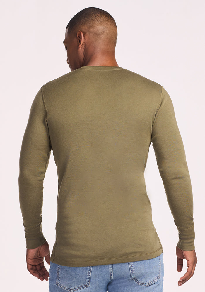 A man stands with his back to the camera, wearing the Woolx Explorer Base Layer in Winter Moss and light blue jeans. The plain, light backdrop emphasizes the comfort and versatility of this everyday essential.