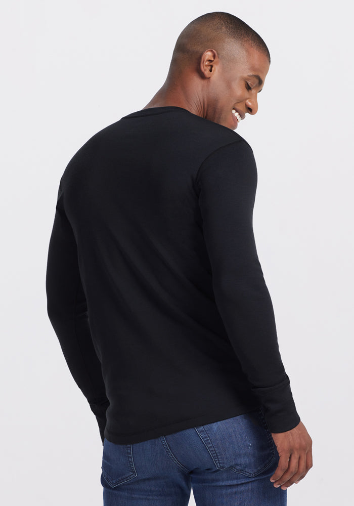 A man smiles over his shoulder, wearing the Woolx Glacier Long Sleeve Crew in black and blue jeans, set against a plain light background.