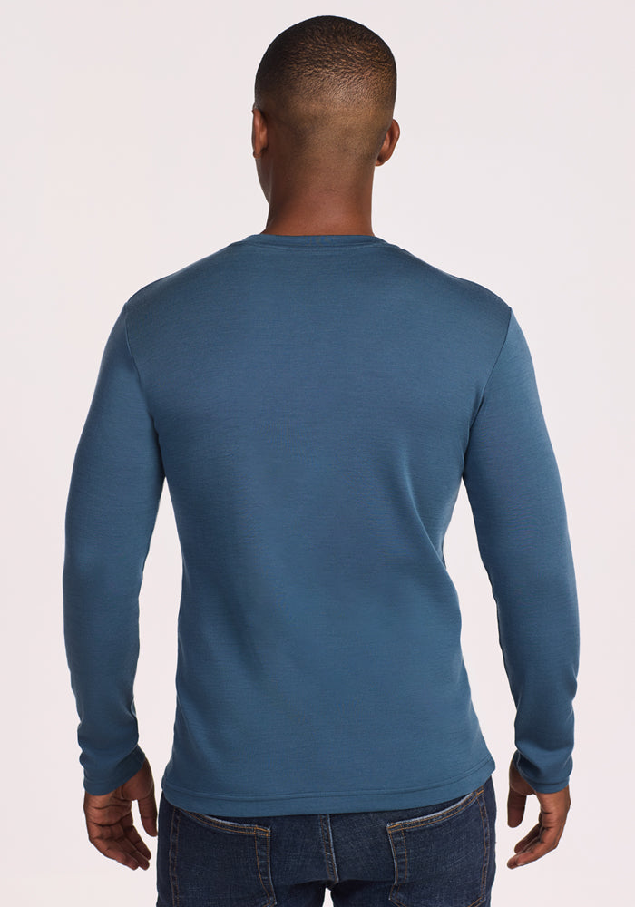 A man faces away from the camera, wearing the Woolx Easton Crew Neck Shirt in Steel Blue—a moisture-wicking merino wool base layer—and dark blue jeans against a plain, light background.