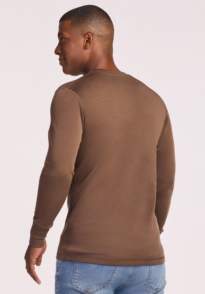 A man wearing the Woolx Explorer Base Layer - Bourbon Oak and light blue jeans stands with his back to the camera, looking over his left shoulder against a light background.