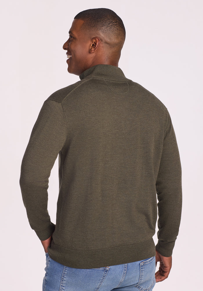 A man faces away, smiling with hands in pockets, wearing the Woolx Enzo Quarter Zip in Pine Needle and light blue jeans. The background is plain and light-colored.