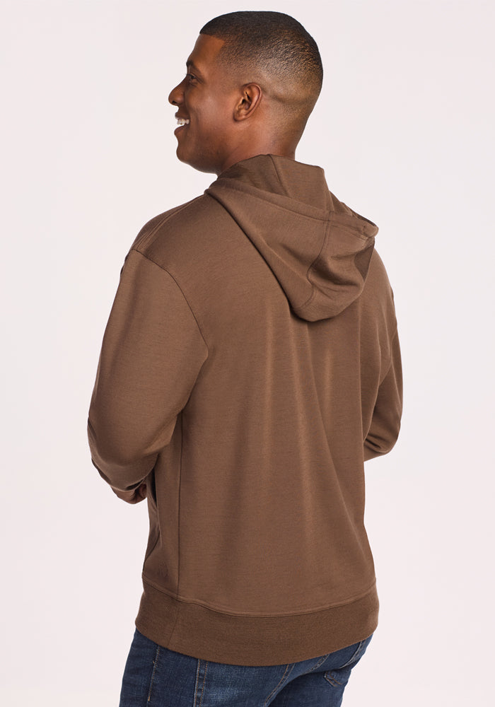 A man, smiling and facing away, wears the Woolx Chase Hoodie in Bourbon Oak with blue jeans against a plain light background.