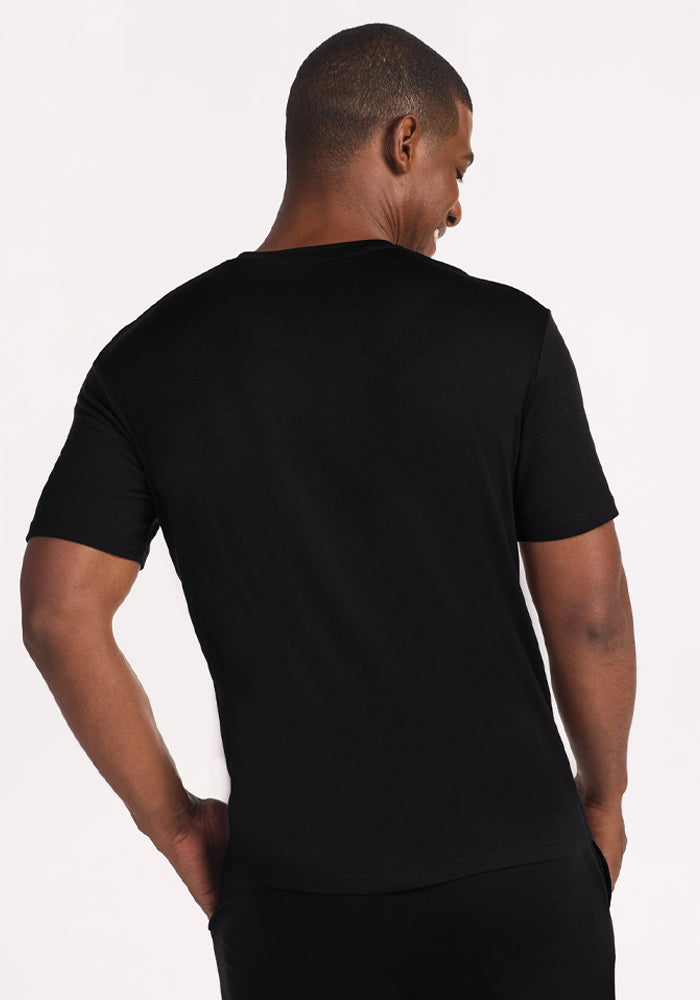 A man stands with his back to the camera, wearing Woolx's Finn Top in black and black pants against a light background.