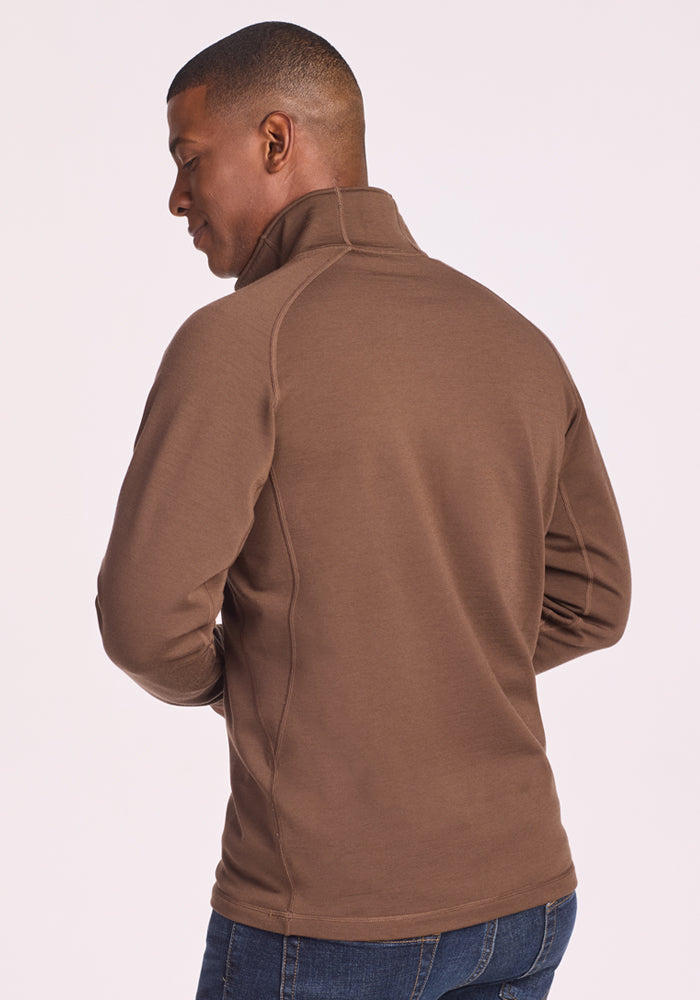 A man stands with his back to the camera, wearing the Woolx Blizzard 1/4 Zip in Bourbon Oak and blue jeans against a plain light background. His head is turned slightly left, and his arms rest relaxed at his sides.