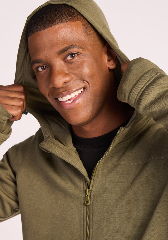 A smiling man in the Woolx Grizzly Sweatshirt in Winter Moss pulls up the hood, looking at the camera against a plain light background.