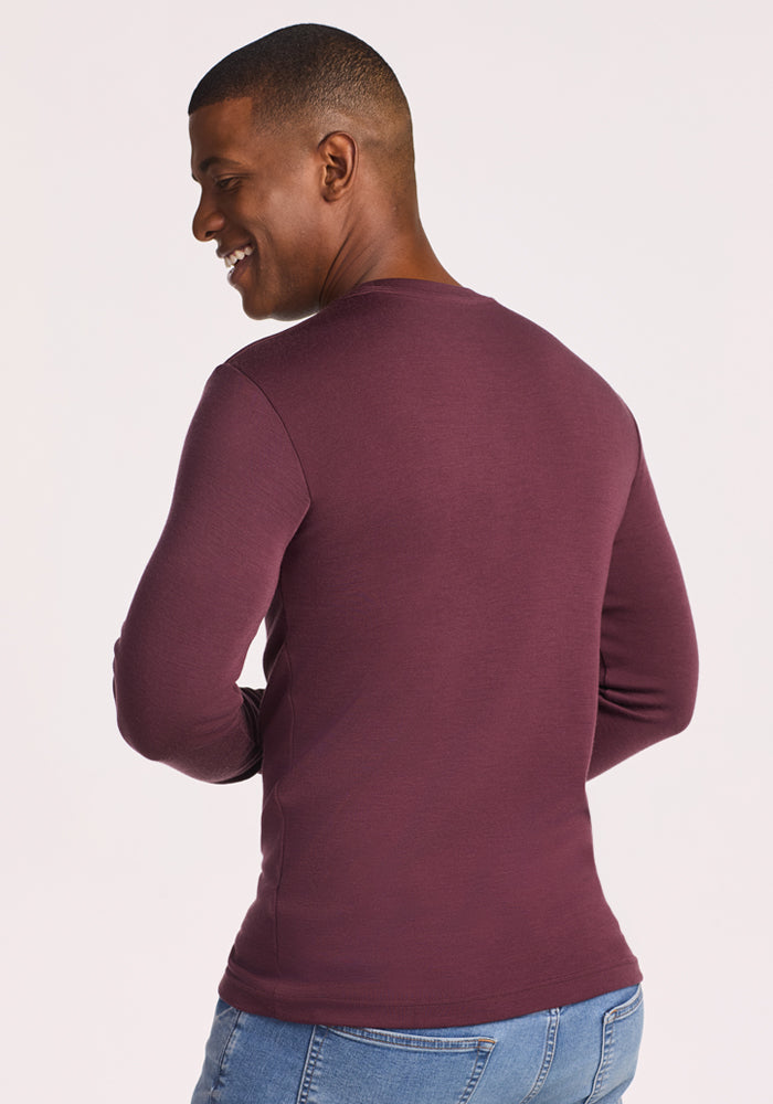 A man with short hair wears the Woolx Easton Crew Neck Shirt in Grape Harvest and blue jeans, standing slightly turned to the side and smiling over his shoulder against a light background.