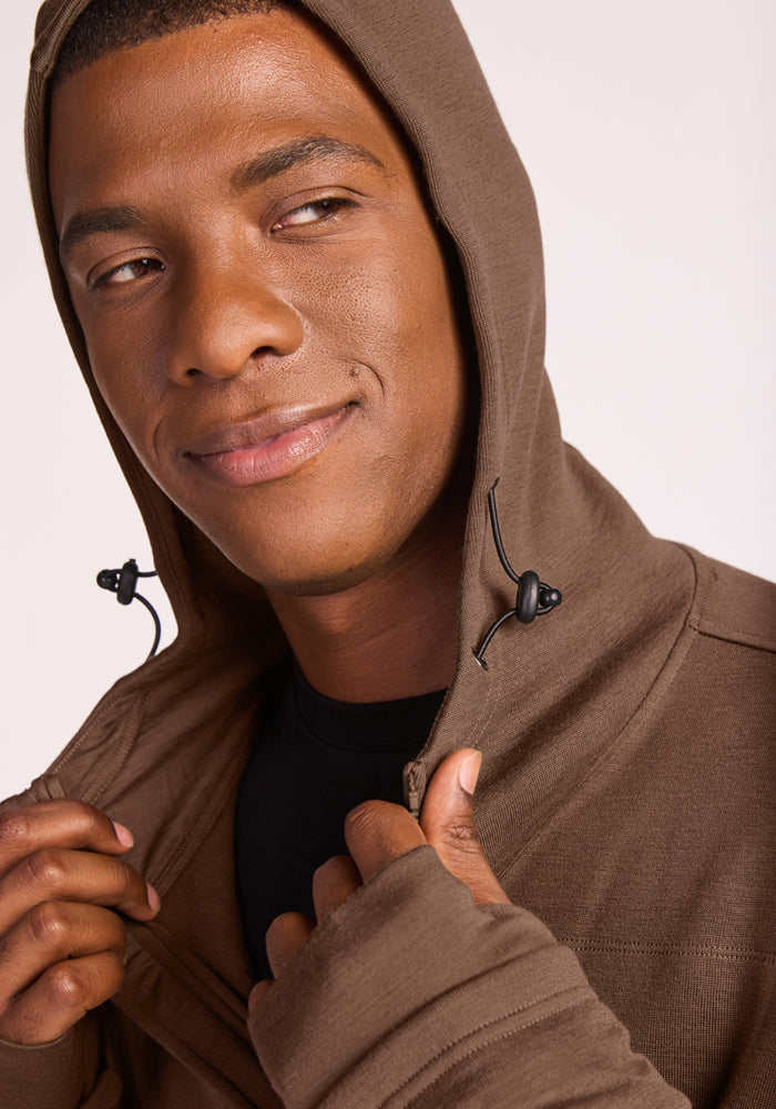 A person wearing the Woolx Grizzly Sweatshirt in Bourbon Oak, featuring black drawstrings, smiles slightly while holding the moisture-wicking Merino wool fabric and zipper near their face.