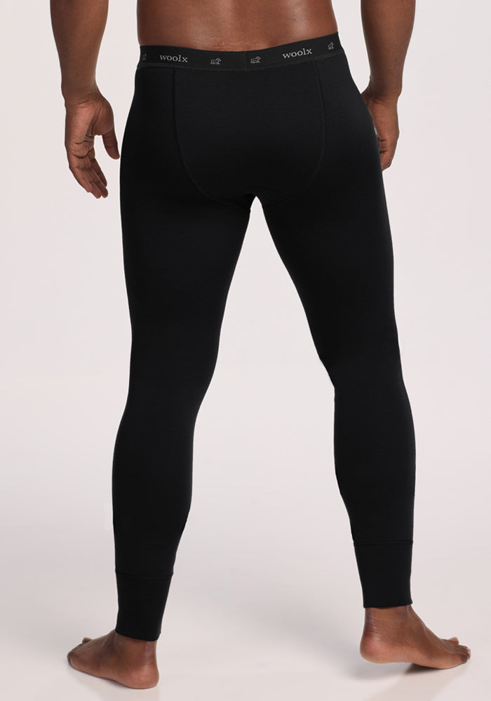 A barefoot person, seen from behind, wears fitted black Woolx Arctic Leggings with a visible branded waistband. The plain light background emphasizes these ultra-heavyweight Merino wool leggings as the ideal base layer.