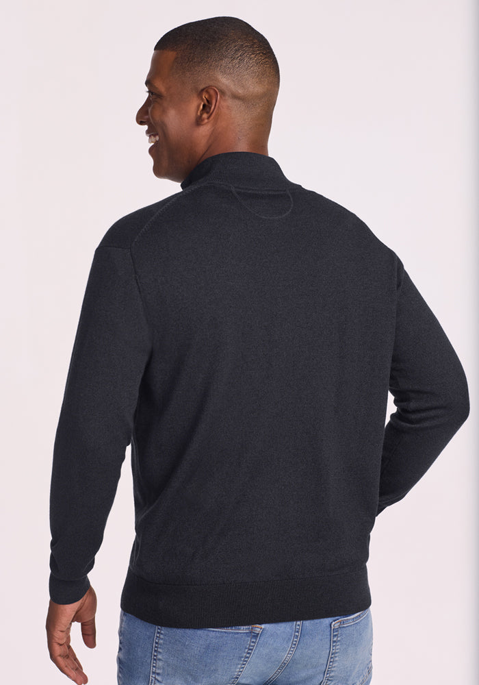 A man with short hair, smiling and facing away, wears the Woolx Enzo Quarter Zip - Coal Stone sweater with light blue jeans. The background is plain and light-colored.